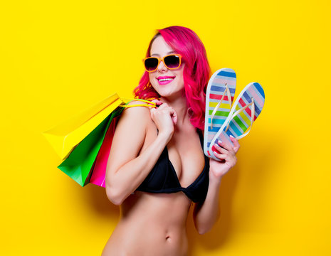 Young pink hair girl in bikini and orange glasses with flip flops and shopping bags. Portrait isolated on yellow background