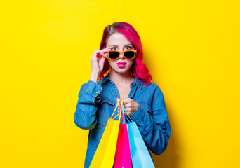 Young pink hair girl in sunglasses and blue shirt holding a colored shopping bags. Portrait on isolated yellow background