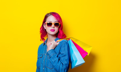 Young pink hair girl in sunglasses and blue shirt holding a colored shopping bags. Portrait on isolated yellow background