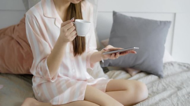 waking up brunette girl is drinking coffee in her bed in morning time in holidays and typing on her smartphone