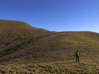 A male hiker in a hat holding trekking poles and walking on a trail. Background is the beautiful alpine landscape of Taiwan with a clear blue sky.