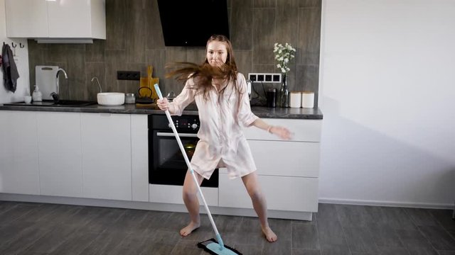 funny girl is having fun, when doing house cleaning, dancing extravagantly around broom in kitchen
