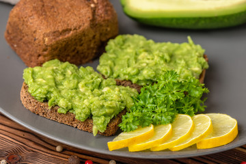 sandwiches with avocado green paste of mashed and cutted toast decorated alligator pear fruit, black bread, sliced lemon on plate on wooden background, healthy eating