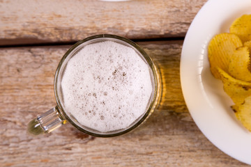 On the wooden table is a mug with a light beer. Nearby is a plate with chips. View from above. Horizontal image.