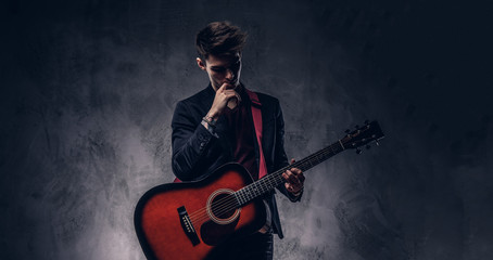 Handsome young thoughtful musician with stylish hair in elegant clothes posing with a guitar in his hands.