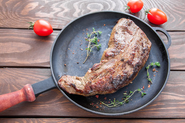 roast beef steak with appetizing crust sprinkled with herbs and spices and twigs of thyme on a black iron frying pan on a brown wooden table with cherry tomatoes top view