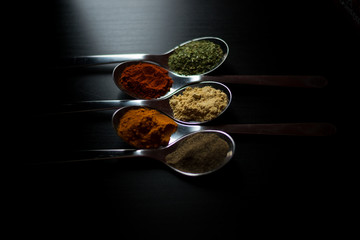 Spices colored in silver spoons, placed on a black wooden table. Pepper, pepper, salt, basil, mustard