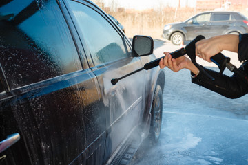 Girl washes a black SUV with a high-pressure car wash. Soap foam and water on the machine. Self-service car wash. A young woman washes the car in the street car wash in the fresh air.