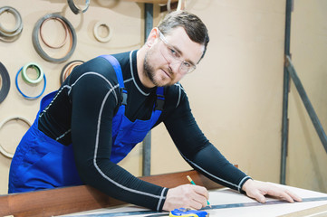 A bearded tired handsome carpenter on a table with a pencil draws a sign on the board. At the...