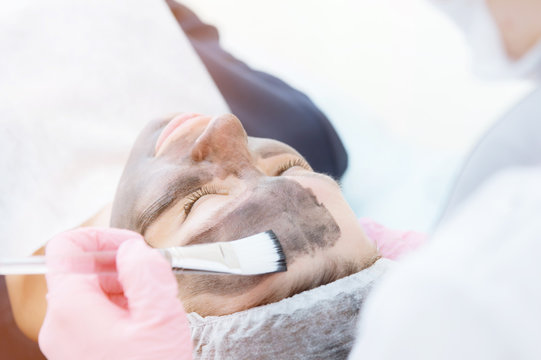The cosmetologist in pink gloves with a brush applies a carbon mask for peeling on the face of a young girl in a cosmetology room. The concept of cosmetology services and self-care. The concept of