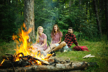 Happy friends having picnic in woods. Cheerful youngsters enjoying summer evening near campfire, weekend in countryside