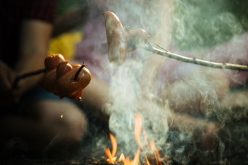 Overnight camping in forest. Cooking sausages on sticks over flames of campfire. Tasty, smoked meat fried on fire