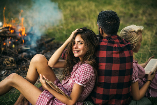 Smiling brunette with braces leaning on her friend. Turn back guy looking at fire while two beautiful girls read books. Relaxation in nature, friendship concept