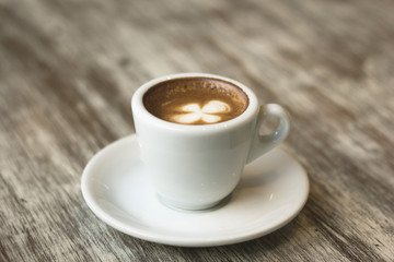 Clover leaf foam shape on top of coffee in white cup with plate on brown wooden table. Vintage effect