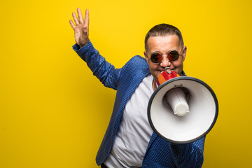 Mature man in sunglasses speaking with loudspeaker isolated on yellow background