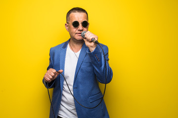 Portrai of mature middle age man in suit singing over the microphone isolated on yellow background. Singer concept.