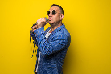 Portrai of mature middle age man in suit singing over the microphone isolated on yellow background....