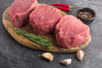 Raw beef meat on a cutting board with rosemary, spices, garlic and chili pepper. Gray stone background