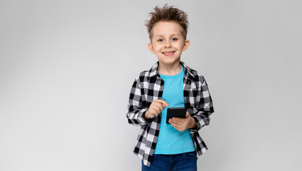 A handsome boy in a plaid shirt, blue shirt and jeans stands on a gray background. The boy is holding a phone
