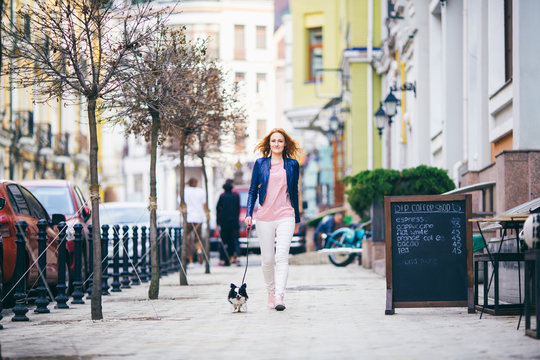 young redhaired Caucasian woman walking along European street with small Chihuahua breed dog of two colors on leash. Cloudy, warm autumn spring weather. Girl Dressed in leather jacket and pink shoes