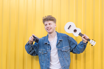 Happy young hipster stands on the background of a yellow wall with ukulele and a retro camera in his hands.