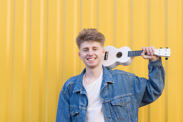 Happy young musician standing with a white ukulele on a yellow background and smiling. Musical concept. Stylish young man with ukulele