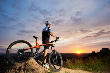 Fototapeta na wymiar Rear view of a guy with a mountain bike standing on a hill in the background of a sunset, a sitting sun and a city in the distance under a beautiful blue sky