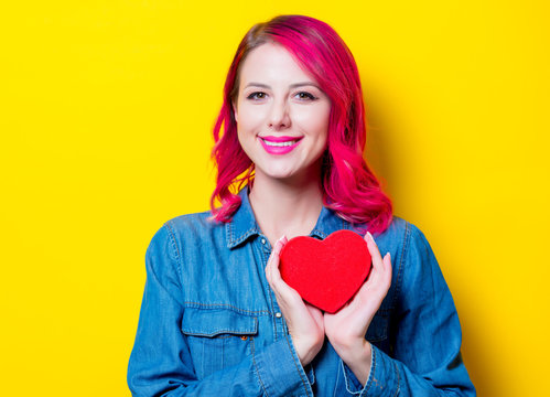 Young pink hair girl in blue shirt holding a red heart shape box. Portrait on isolated yellow background