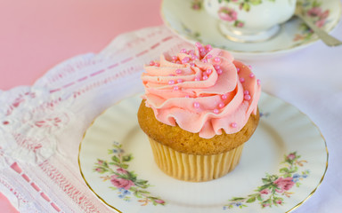 Pink glitter and pearl frosted cup cake on plate with teacup in background - space for text