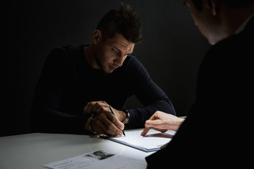 Criminal man with handcuffs signing document in interrogation room - obrazy, fototapety, plakaty