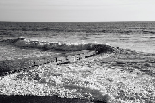 Fototapeta Zigzag of a sea wave breaking ashore and breakwater with sea foam, black and white photography