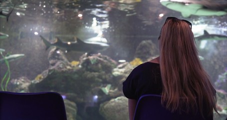  The girl is looking at a large aquarium. The girl looks at the fish in the aquarium.