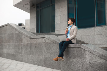 Time for having a rest! Dreaming smart casual young man sitting outdoor and drinking coffee-to-go, while being at the airport and waiting his flight.