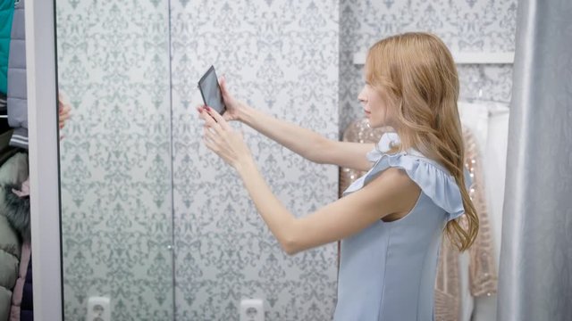 young woman is taking photo of her reflection in a mirror of fitting room of clothing store, using smartphone