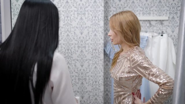 blonde girl is trying a shiny dress in a fitting room , looking in mirror and advising with her brunette friend