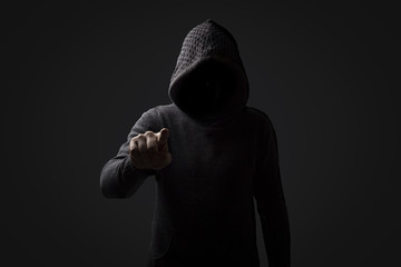 Faceless man in a hood with points a finger at the viewer on a dark background. Concept Threats and...