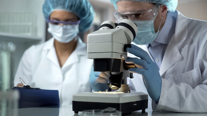 Researcher viewing sample under microscope, assistant taking notes, laboratory