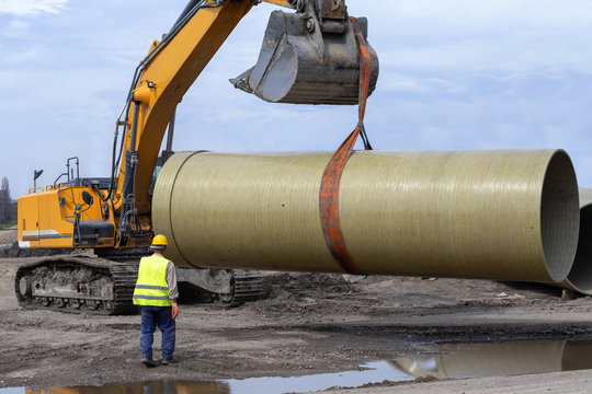 Excavator Bucket Lifting Large Underground Pipe With Worker Helping