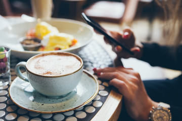 Close-up of woman's hands holding cell phone while sitting in modern cafe and having breakfasts. Lifestyle, reading news, social network, surfing the web / internet in smartphone.