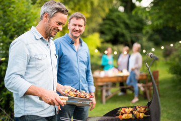 in a summer evening,  two men  in their forties prepares a barbecue for  friends gathered around a table in the garden - obrazy, fototapety, plakaty