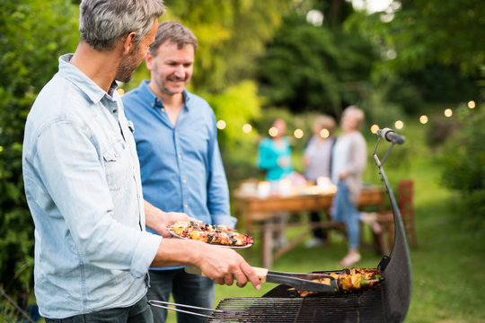 In A Summer Evening,  Two Men  In Their Forties Prepares A Barbecue For  Friends Gathered Around A Table In The Garden