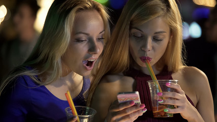 Pretty girls drinking cocktails at bar, checking social networks on smartphone