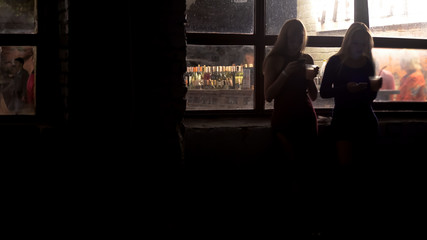 Two beautiful women drinking cocktails, browsing on smartphones at nightclub