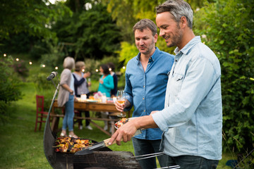 in a summer evening,  two men  in their forties prepares a barbecue for  friends gathered around a table in the garden