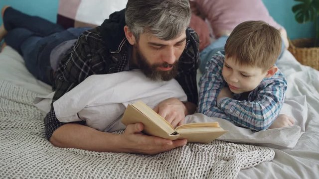 Loving parent bearded man is reading interesting book to his curious little son on double bed at home. Parenthood, happy people, love and family concept.