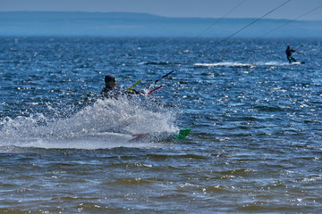 Fototapeta premium A male kiteboarder rides on a board on a large river. He performs various exercises while moving on water. Splashes of water scatter in different directions. The sun's rays shine in the water. 