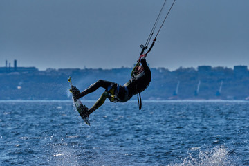 A male kiteboarder rides on a board on a large river. He performs various exercises while moving on water. Splashes of water scatter in different directions. The sun's rays shine in the water. 