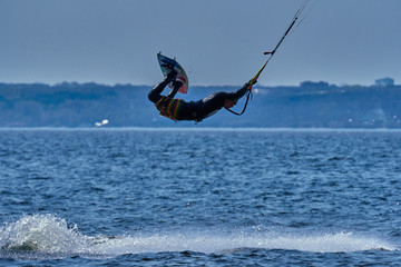 Naklejka premium A male kiteboarder rides on a board on a large river. He performs various exercises while moving on water. Splashes of water scatter in different directions. The sun's rays shine in the water. 