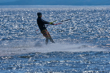 Fototapeta premium A male kiteboarder rides on a board on a large river. He performs various exercises while moving on water. Splashes of water scatter in different directions. The sun's rays shine in the water. 