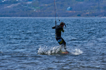 Obraz premium A male kiteboarder rides on a board on a large river. He performs various exercises while moving on water. Splashes of water scatter in different directions. The sun's rays shine in the water. 